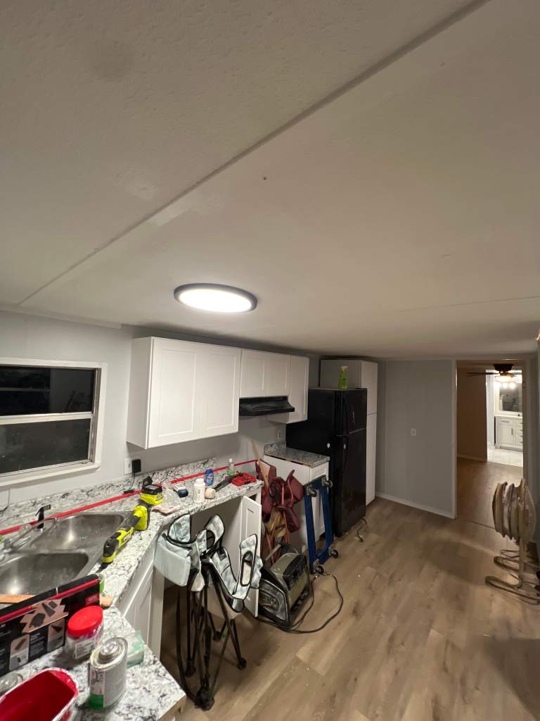 Interior of a kitchen undergoing renovation, featuring a granite countertop, modern white cabinetry, and a large window. Construction tools and equipment are visible, indicating ongoing work.